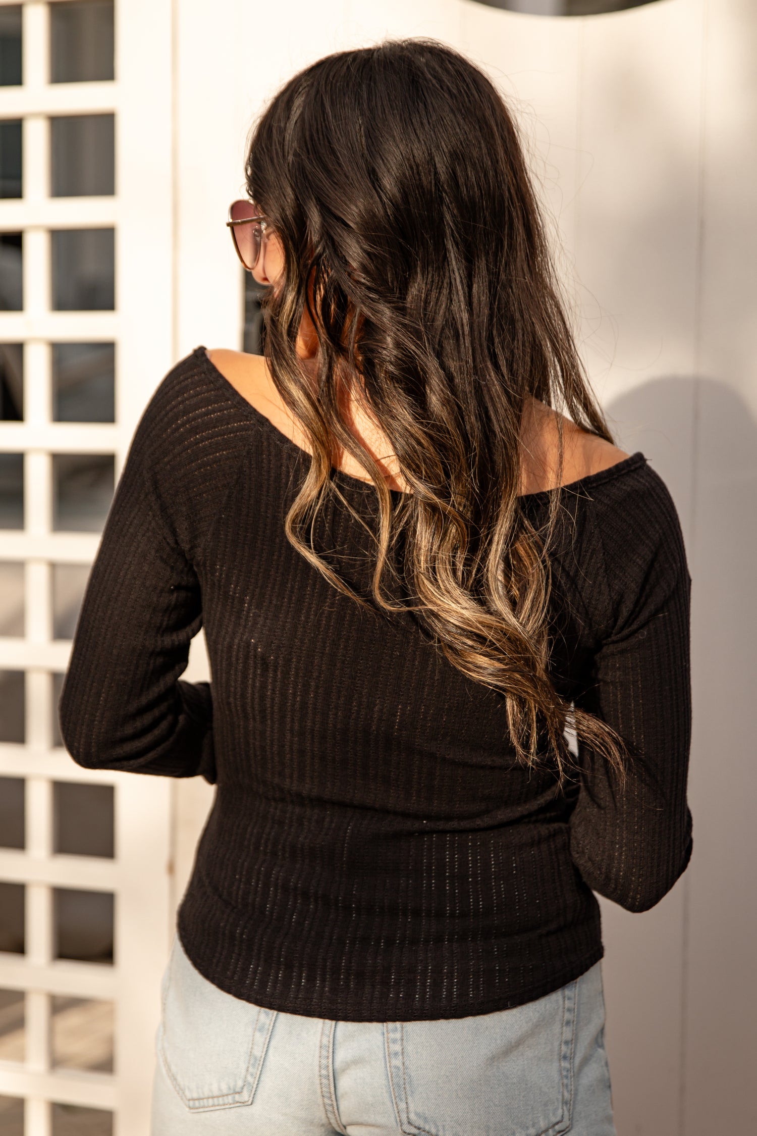 Woman wearing a black long-sleeve top and light blue jeans, standing against a white wall.