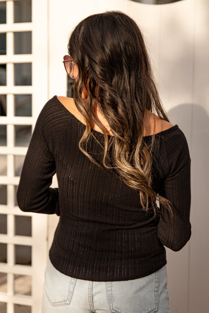 Woman wearing a black long-sleeve top and light blue jeans, standing against a white wall.