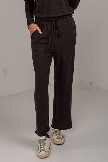 A person wearing dark brown wide-leg drawstring sweatpants with side pockets, paired with a matching dark top and beige sneakers, standing in a studio setting.
