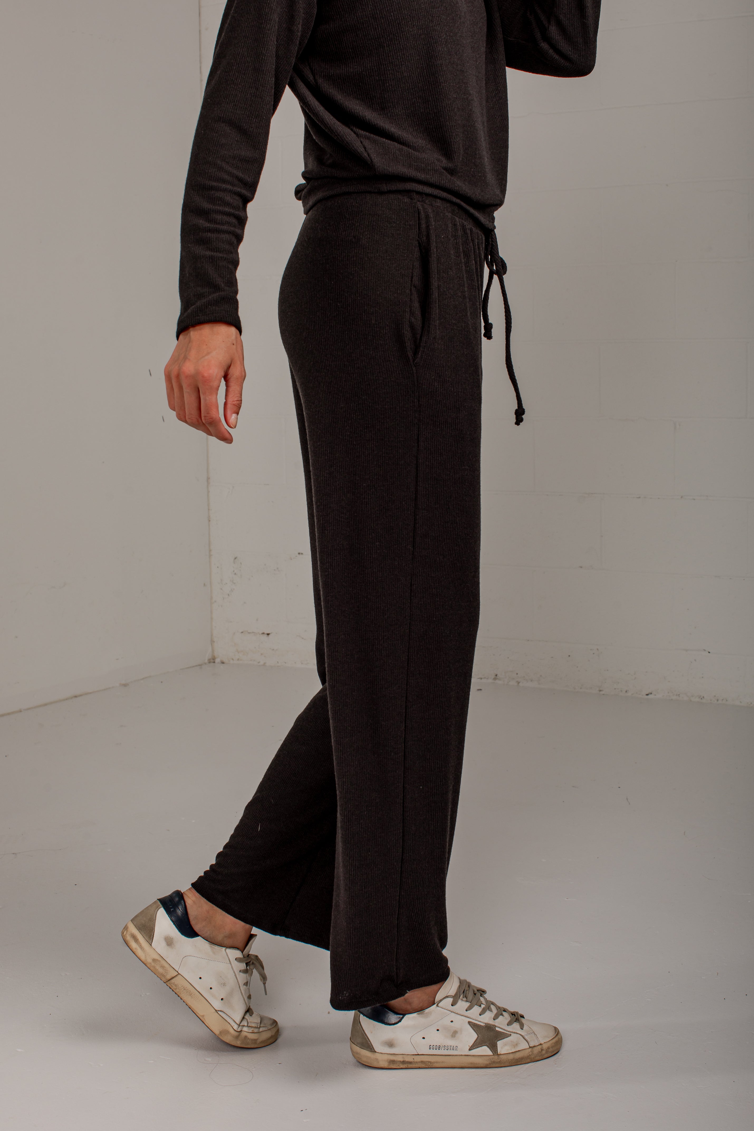 A person wearing dark brown wide-leg sweatpants with a drawstring waist, paired with a matching dark top and white sneakers, standing in a minimalist studio setting.