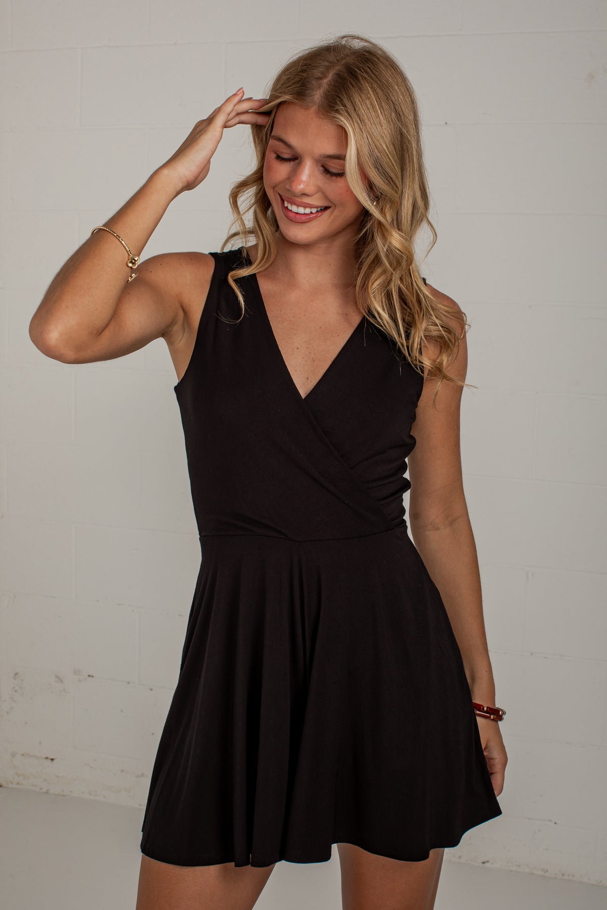 A woman modeling a sleeveless black wrap-style mini dress with a flared skirt, standing against a plain light gray wall.