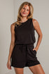 A woman modeling a black sleeveless top and matching black shorts with a drawstring waist, standing against a light gray brick wall.