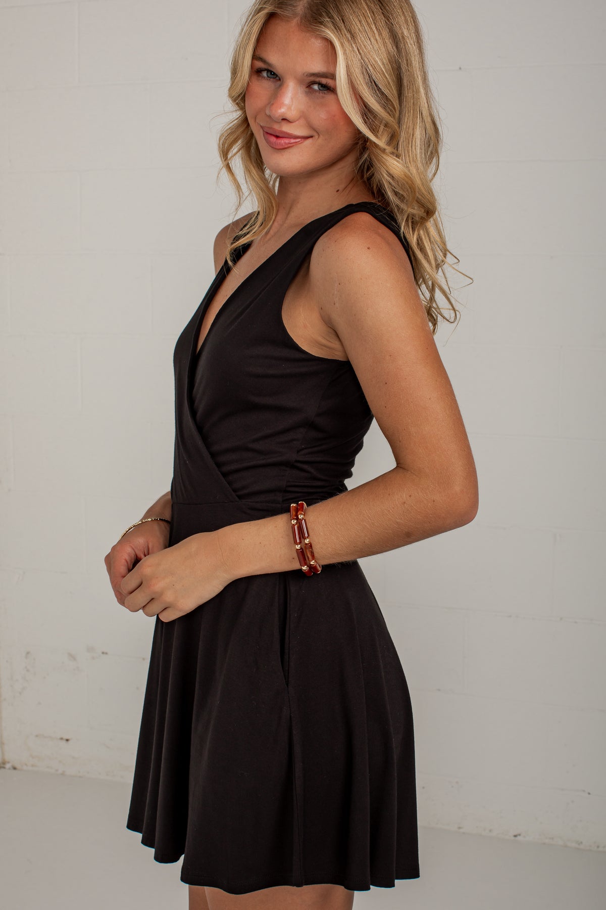 A blonde woman wearing a sleeveless black wrap-style mini dress with a flared skirt, standing against a plain white wall.
