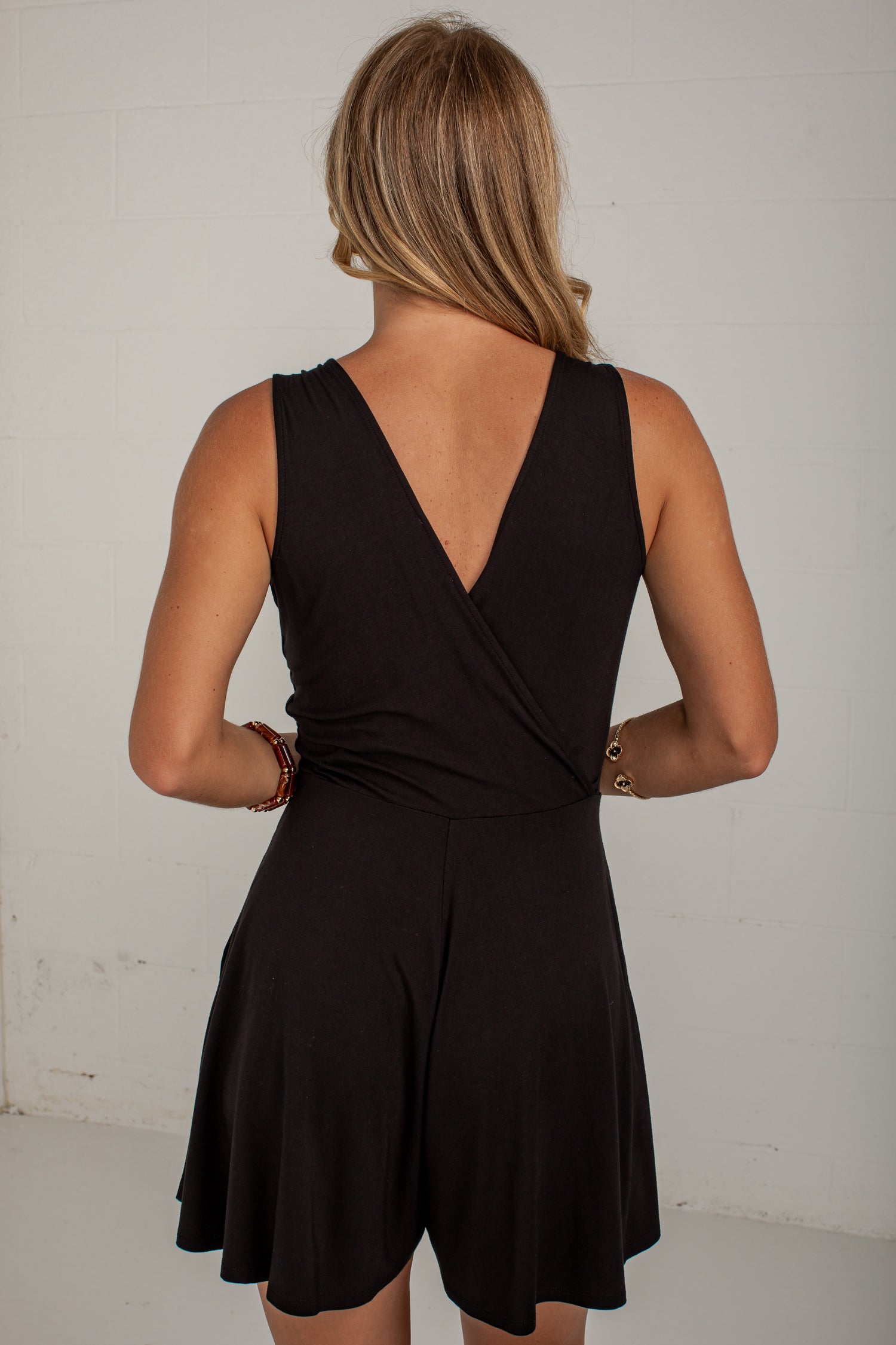 A woman viewed from behind wearing a black sleeveless romper with a wrap-style V back and flared shorts, standing against a plain light wall.