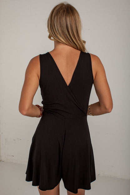 A woman viewed from behind wearing a black sleeveless romper with a wrap-style V back and flared shorts, standing against a plain light wall.