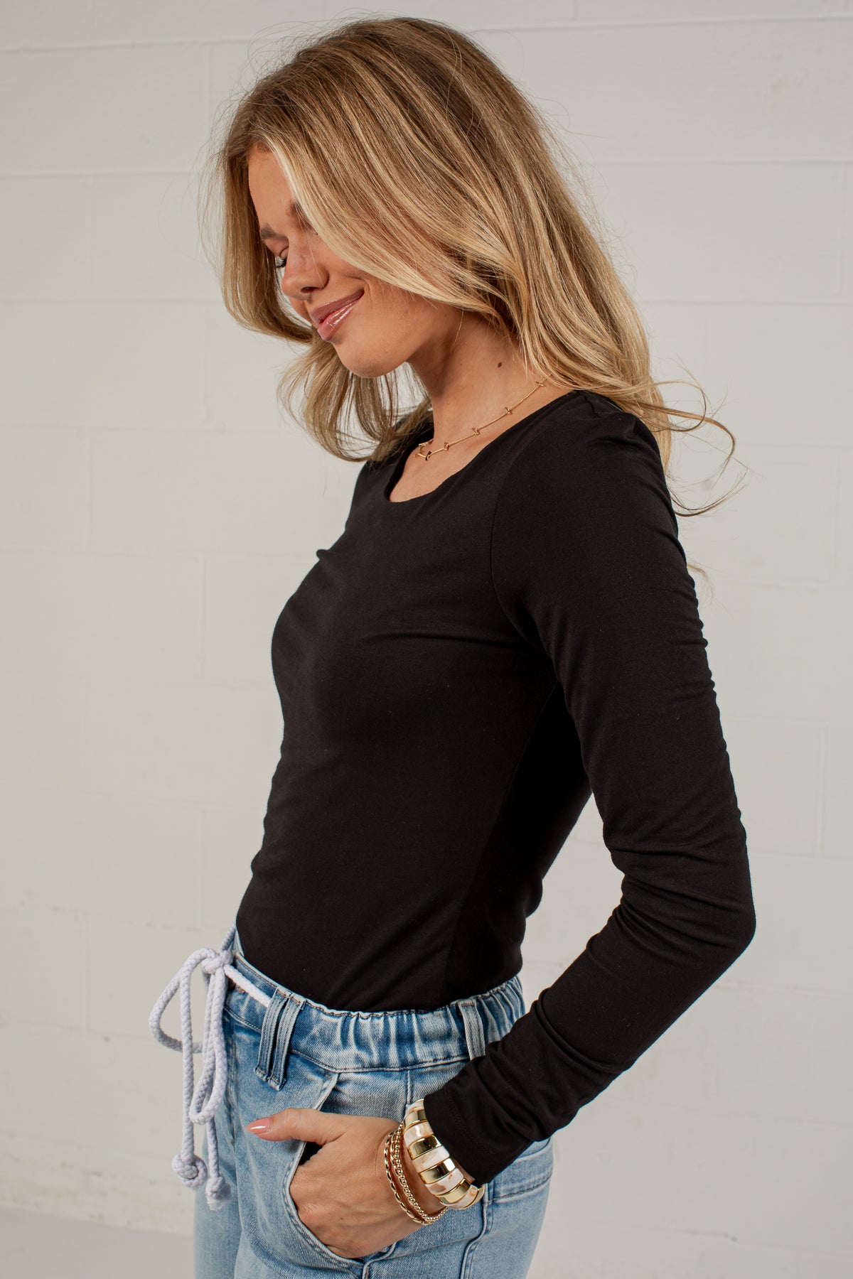 A woman wearing a black long-sleeve scoop neck top and light blue jeans, standing against a white paneled wall.