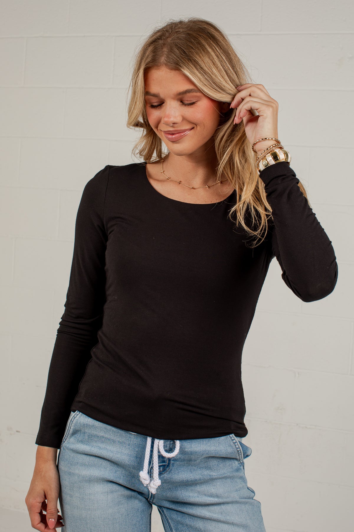 A woman wearing a black long-sleeve crewneck top and light blue jeans, standing against a white brick wall.