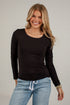 A smiling blonde woman wearing a fitted black long-sleeve scoop-neck top and light blue jeans, standing against a white brick wall.