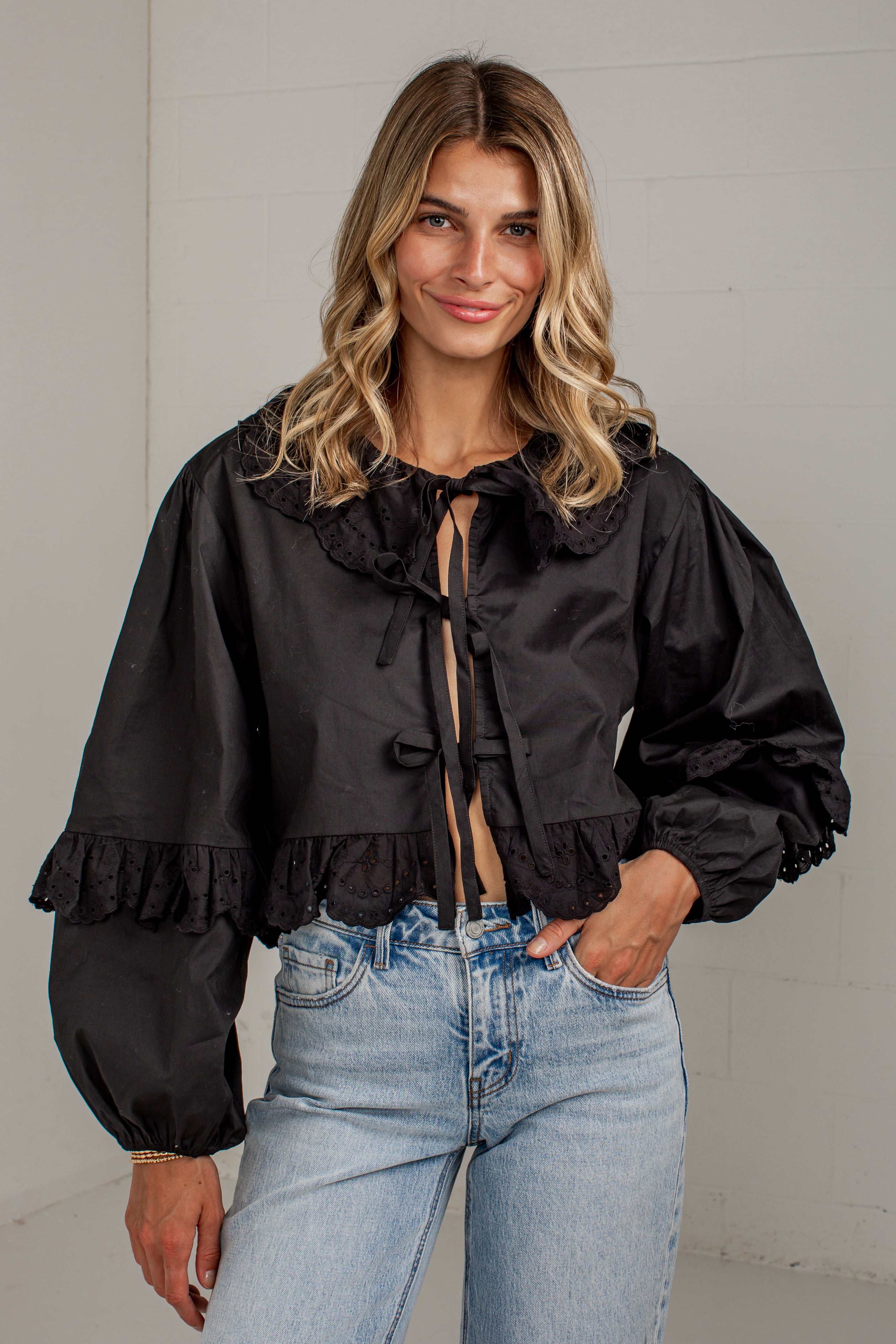 A woman wearing a black long-sleeve blouse with a ruffled collar, front tie detail, and gathered puff sleeves, paired with light blue jeans, standing against a light gray wall.