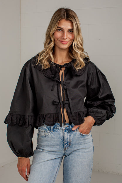 A woman wearing a black long-sleeve blouse with a ruffled collar, front tie detail, and gathered puff sleeves, paired with light blue jeans, standing against a light gray wall.