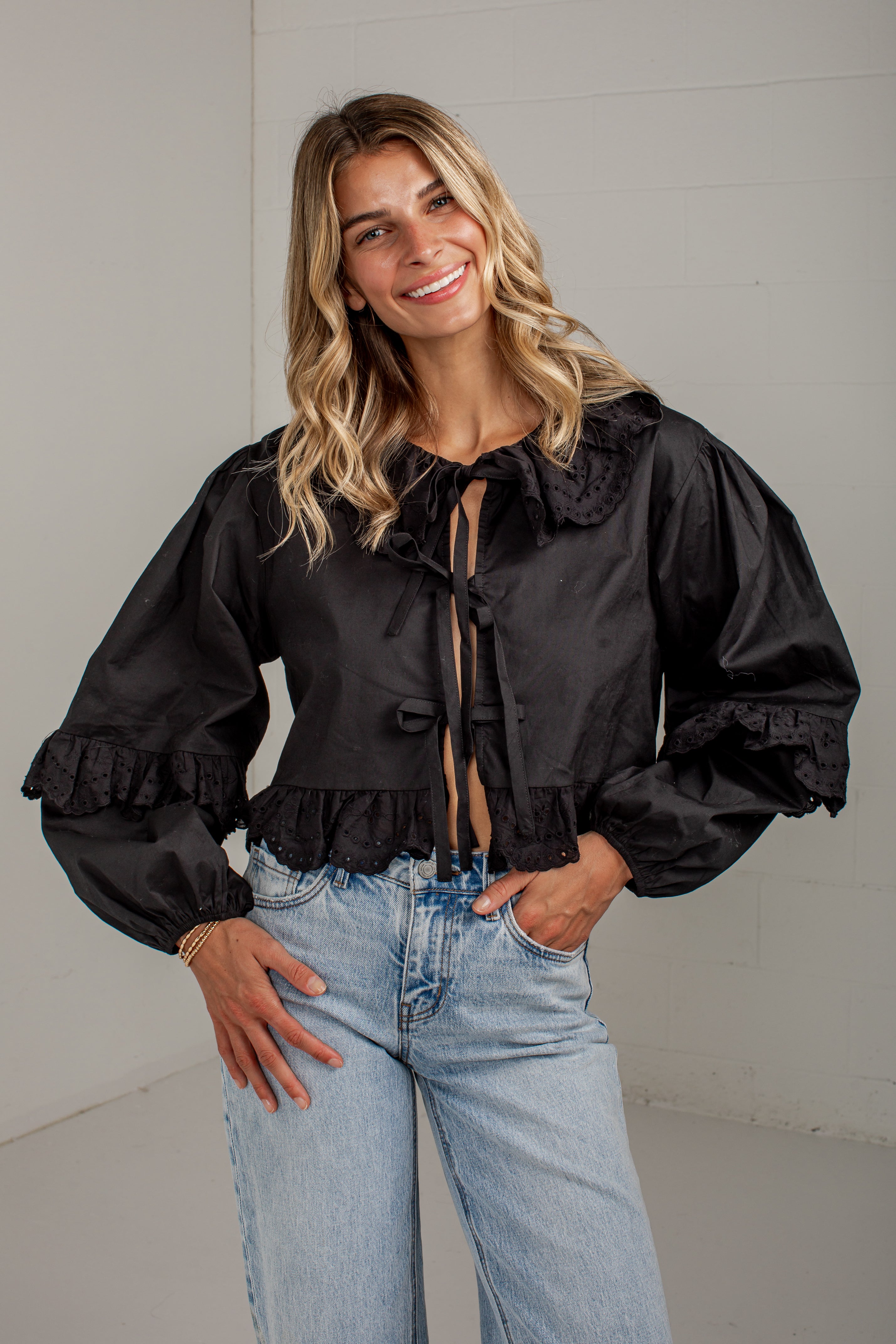 A woman wearing a black long-sleeve cropped blouse with a large ruffled collar, front tie details, and ruffled cuffs, paired with light blue jeans, standing against a light gray wall.
