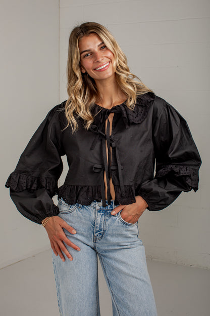 A woman wearing a black long-sleeve cropped blouse with a large ruffled collar, front tie details, and ruffled cuffs, paired with light blue jeans, standing against a light gray wall.