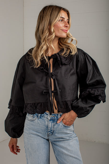 A woman wearing a black cropped satin blouse with dramatic puffed sleeves, ruffled collar and cuffs, and tie-front detailing, paired with light-wash blue jeans against a light gray wall.