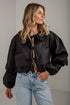 A woman wearing a black cropped satin blouse with dramatic puffed sleeves, ruffled collar and cuffs, and tie-front detailing, paired with light-wash blue jeans against a light gray wall.