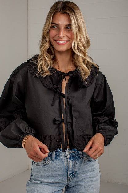 A woman wearing a black long-sleeve blouse with a large ruffled collar and front tie detail, paired with light-wash blue jeans, standing against a light gray paneled wall.