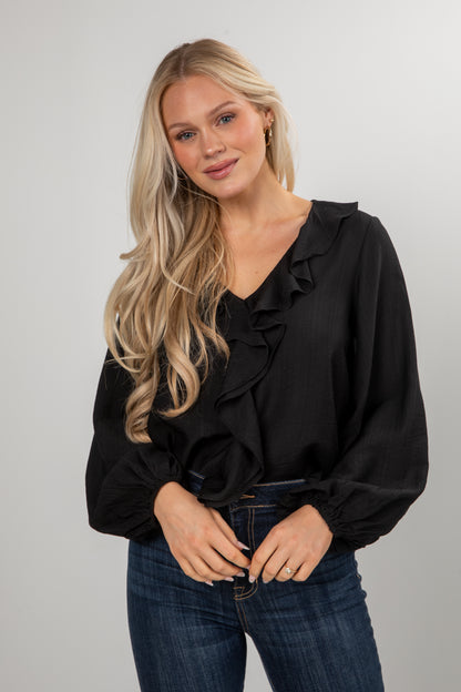 Woman wearing a black blouse with ruffles and blue jeans against a plain background