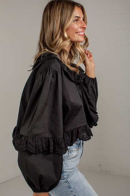 A woman wearing a black long-sleeve blouse with dramatic puff sleeves, lace trim, and gathered ruffles, paired with light-wash blue jeans, photographed against a plain light gray wall.