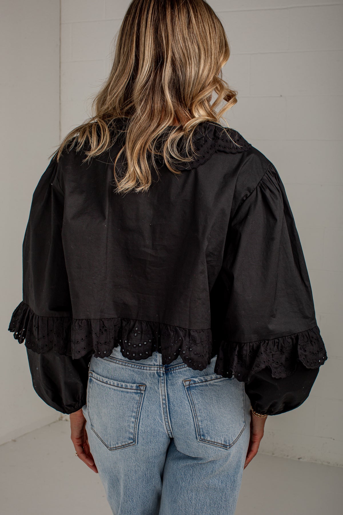 A woman viewed from behind wearing a black cropped blouse with dramatic puffed sleeves, lace trim at the collar and cuffs, and light blue jeans against a white paneled wall.