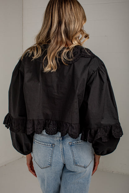 A woman viewed from behind wearing a black cropped blouse with dramatic puffed sleeves, lace trim at the collar and cuffs, and light blue jeans against a white paneled wall.