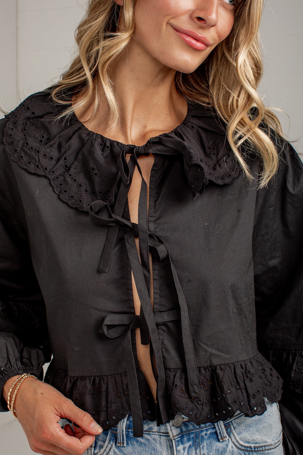 A close-up of a woman wearing a black long-sleeve blouse with a large eyelet lace Peter Pan collar, tie-front keyhole detail, and ruffled hem, paired with light blue jeans.