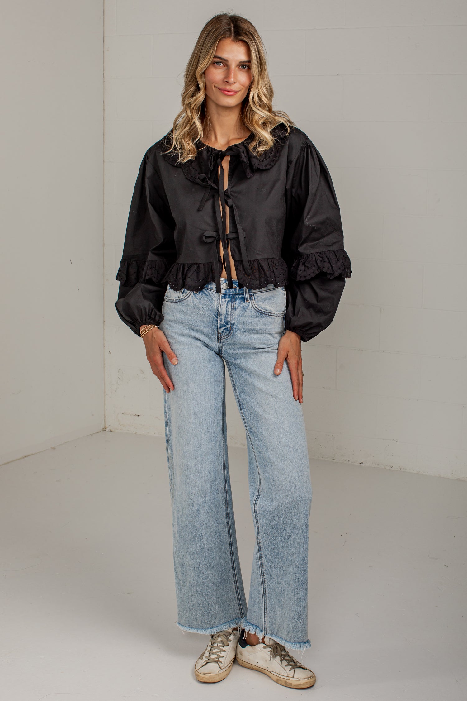 A woman modeling a black long-sleeve blouse with a large ruffled collar and front tie detail, paired with light-wash wide-leg jeans and beige sneakers, standing in a white studio corner.