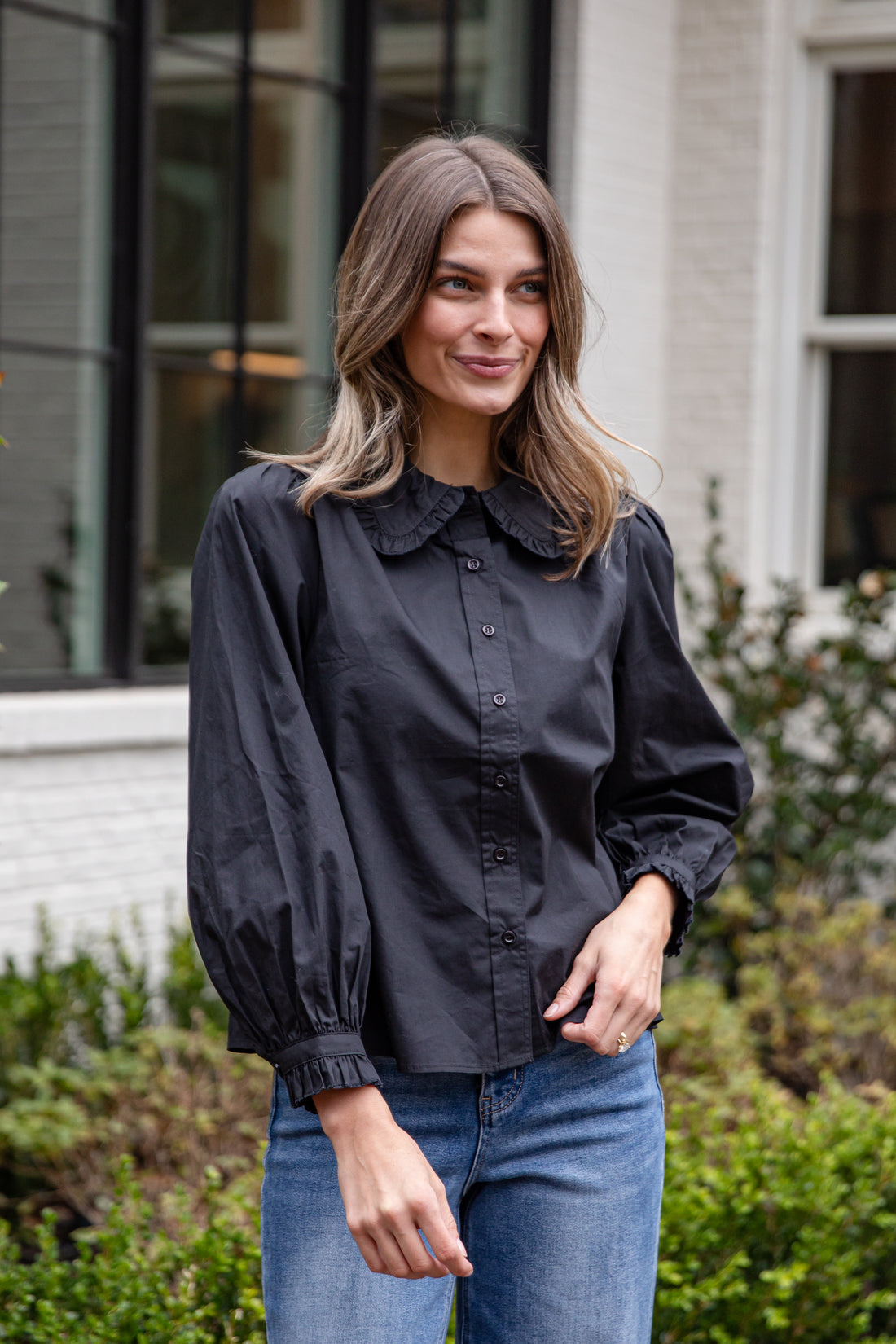 Woman wearing a black blouse with a high collar and blue jeans standing outdoors.