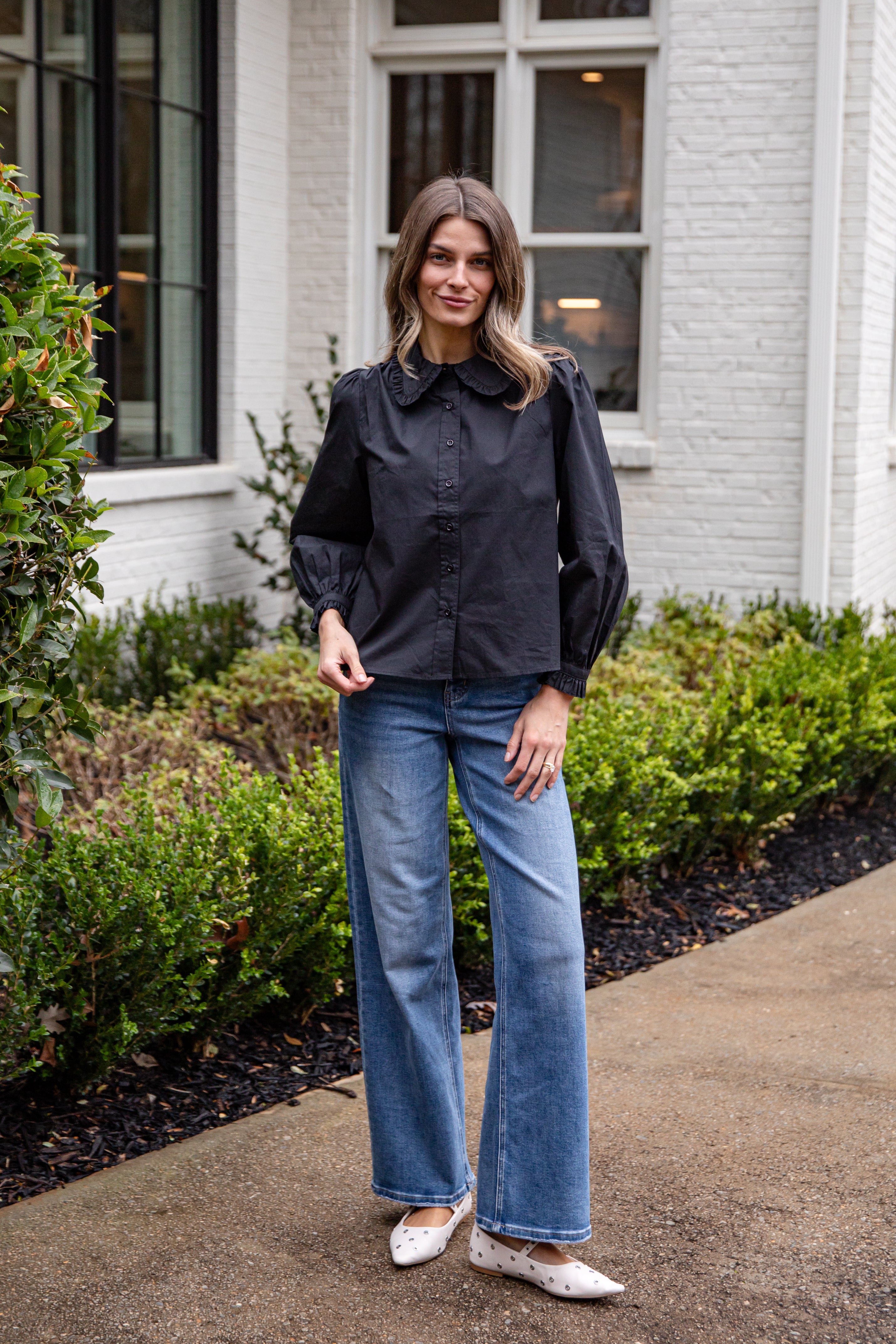 Woman wearing a black top and blue jeans standing outdoors near a building.