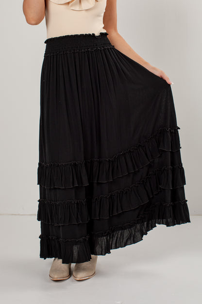 A black tiered maxi skirt with an elastic waistband, shown on a model wearing a cream top and beige boots against a light gray studio background.