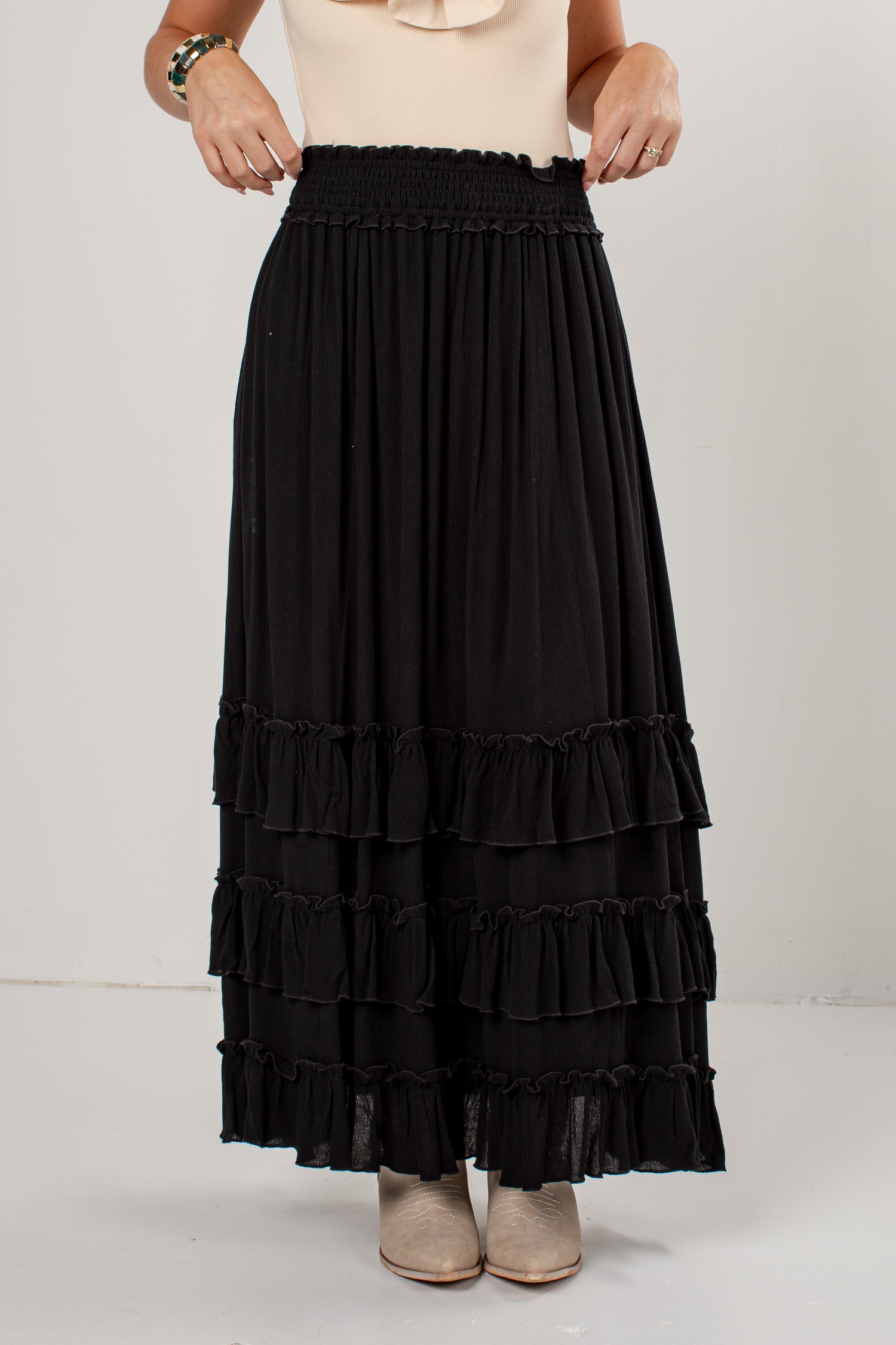A black tiered maxi skirt with an elastic waistband, worn by a model with a beige top and beige shoes, photographed against a plain light gray studio background.