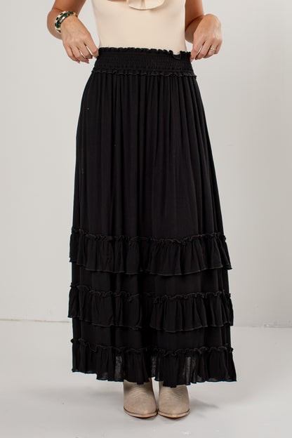 A black tiered maxi skirt with an elastic waistband, worn by a model with a beige top and beige shoes, photographed against a plain light gray studio background.