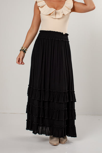 A model wearing a long black tiered maxi skirt with a smocked waistband, paired with a cream ruffled top and beige shoes, shown from the chest down against a plain light gray studio background.