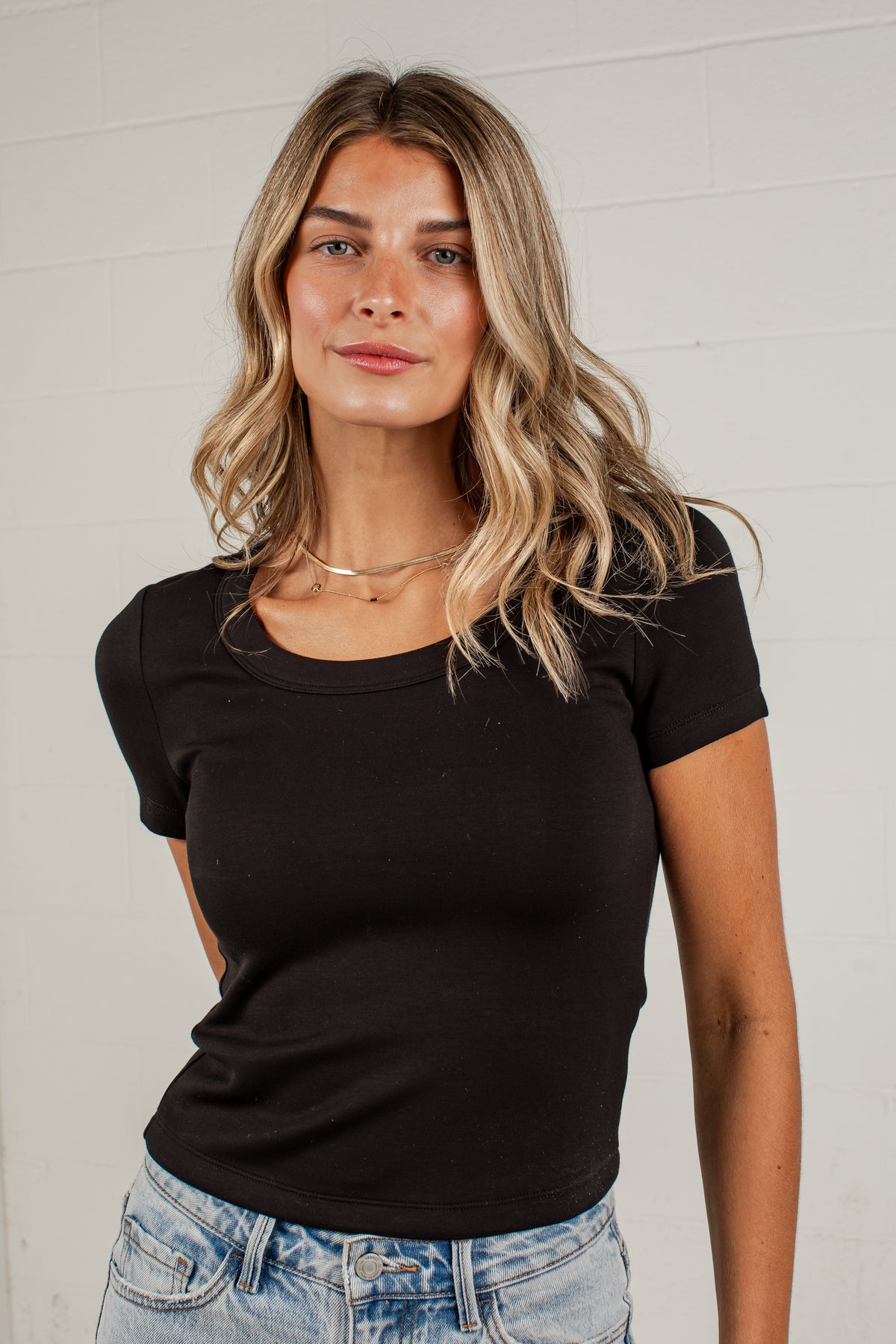 A woman with blonde-highlighted hair wearing a fitted black short-sleeve scoop-neck t-shirt and light-wash blue jeans, standing against a white brick wall.