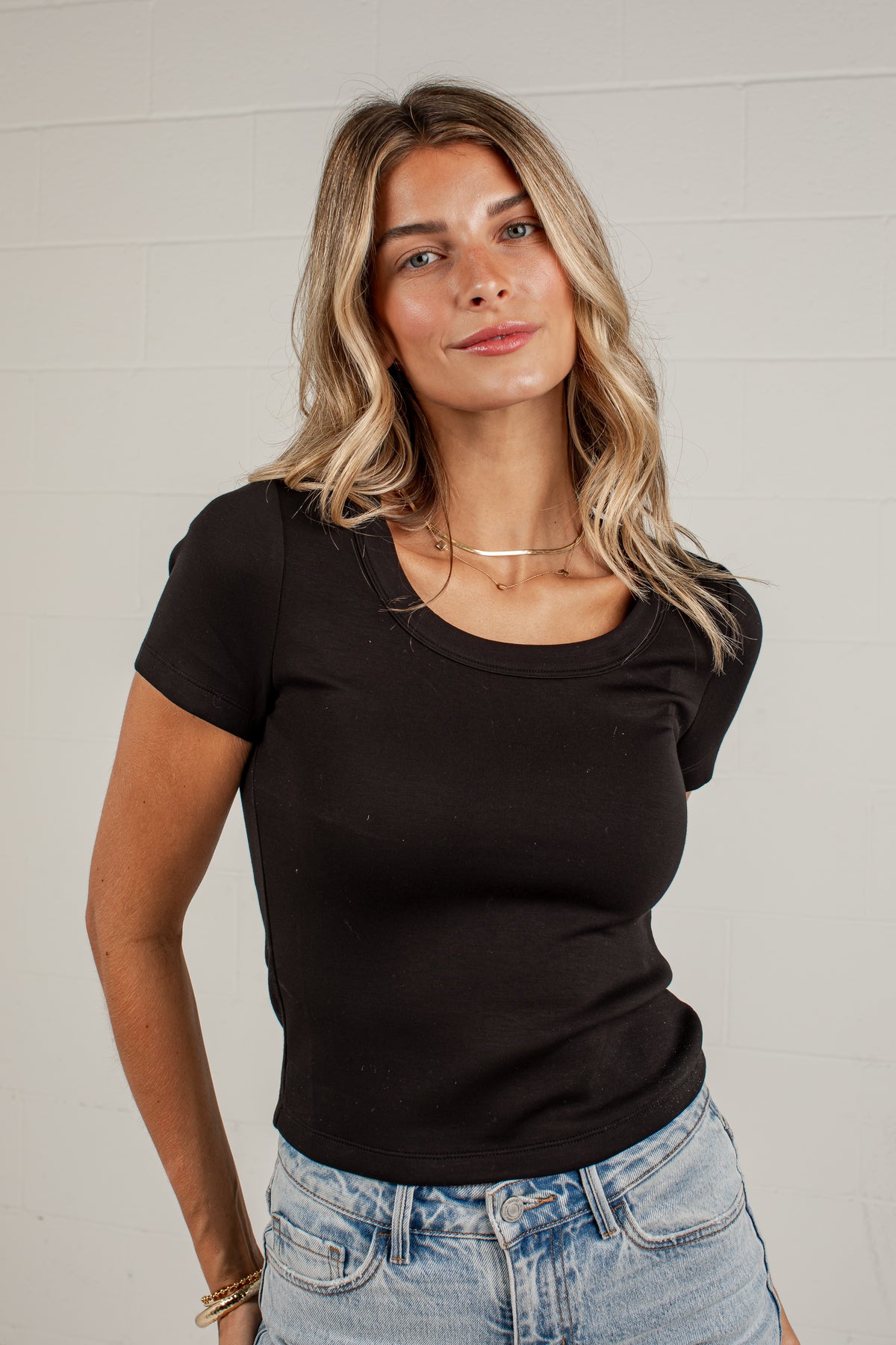 A woman wearing a fitted black short-sleeve scoop-neck t-shirt and light-wash blue jeans, standing against a white brick wall.