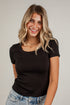 A smiling woman with blonde-highlighted hair wearing a fitted black short-sleeve scoop-neck t-shirt and light-wash blue jeans, standing against a white paneled wall.