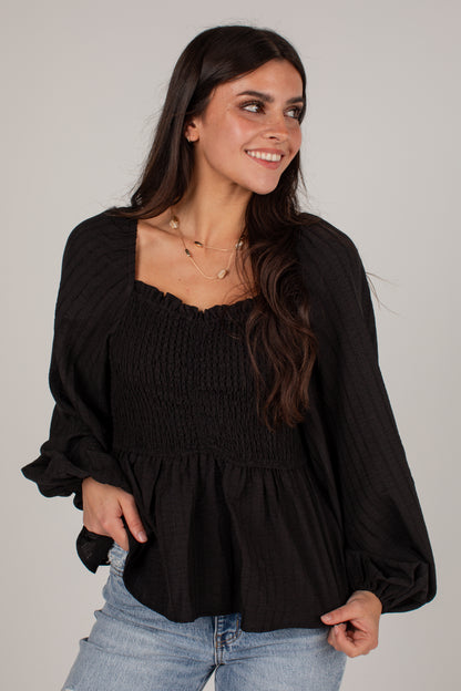 A woman wearing a black long-sleeve smocked blouse with a square neckline and voluminous sleeves, paired with light blue jeans, against a plain light gray studio background.