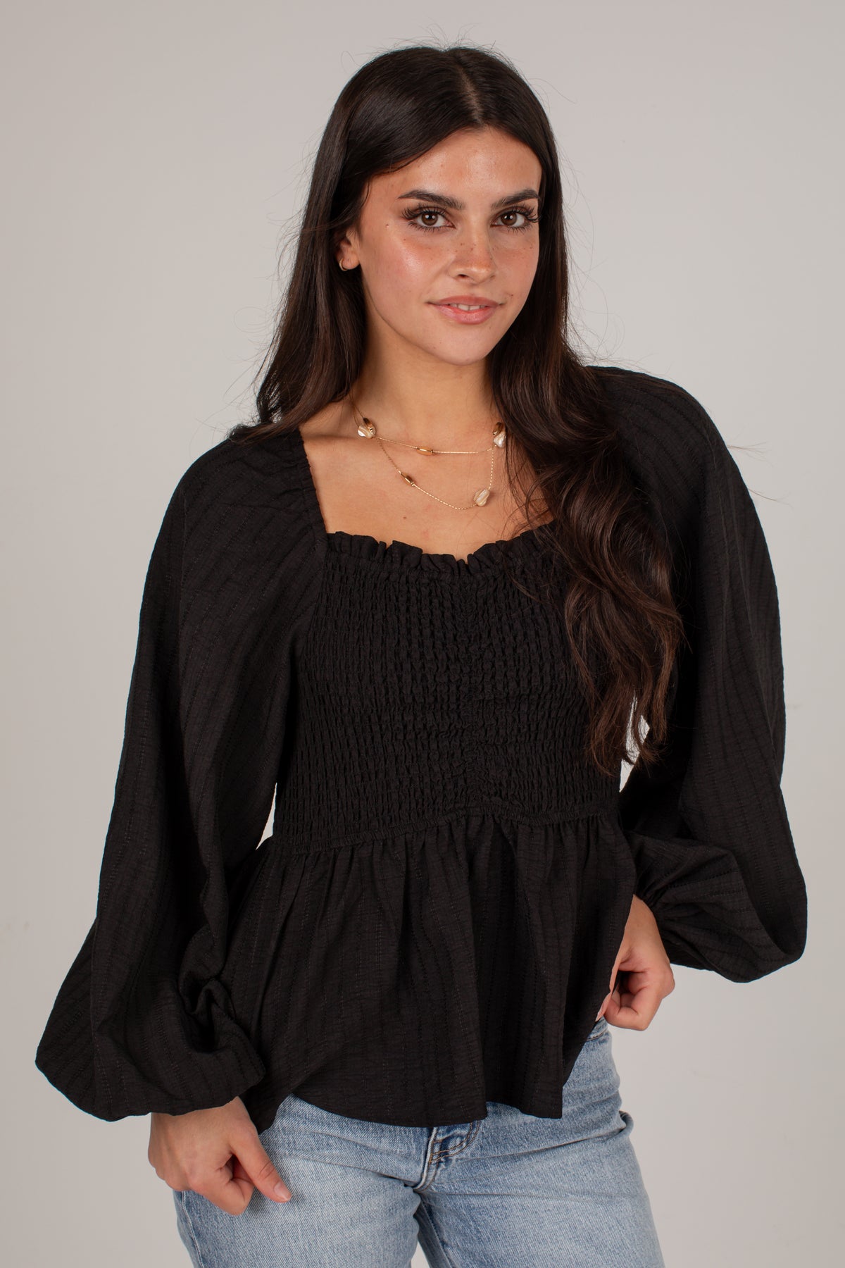 A woman wearing a black long-sleeve smocked blouse with puff sleeves and a square neckline, paired with light blue jeans, against a plain light gray background.