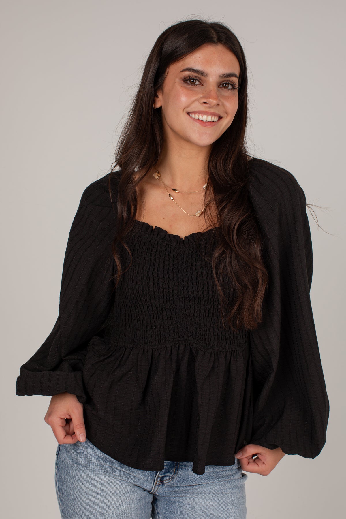 A woman with long dark hair smiling, wearing a black long-sleeve smocked blouse with a peplum hem and light blue jeans, against a plain light gray background.