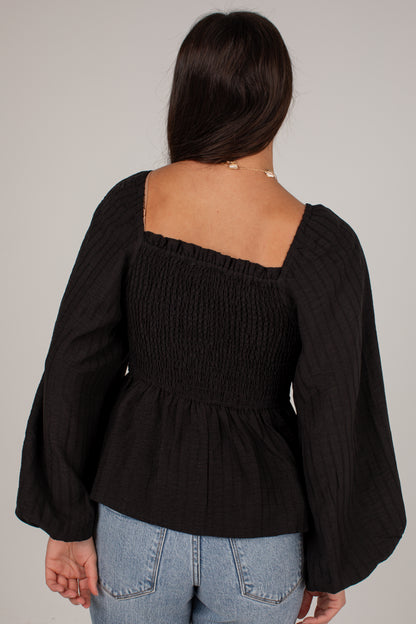A woman shown from the back wearing a black smocked long-sleeve top with a square neckline and flared sleeves, paired with light blue jeans, against a plain light gray background.