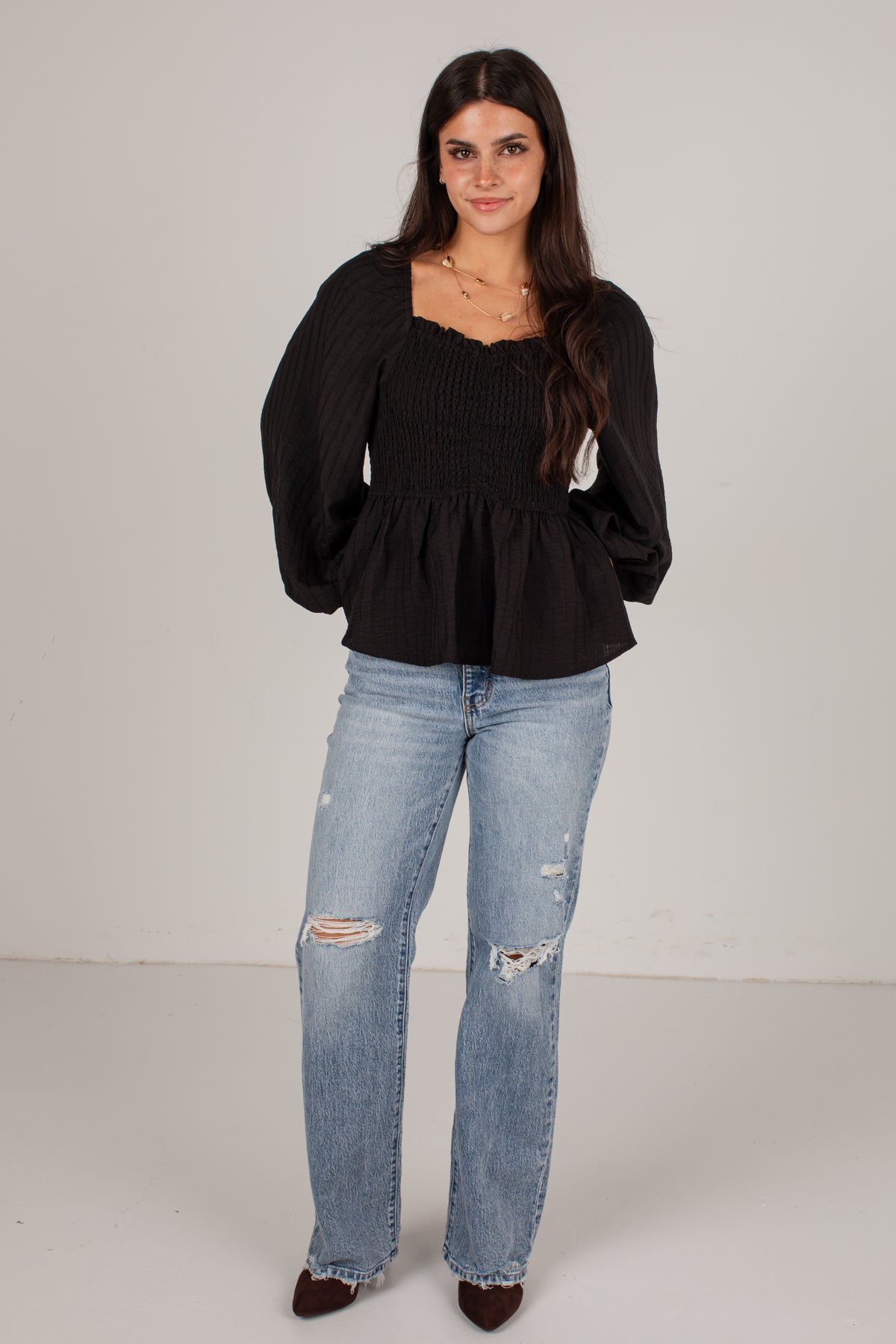 A woman modeling a black long-sleeve smocked peplum top paired with light-wash distressed jeans and brown pointed-toe heels, standing against a plain light gray studio background.