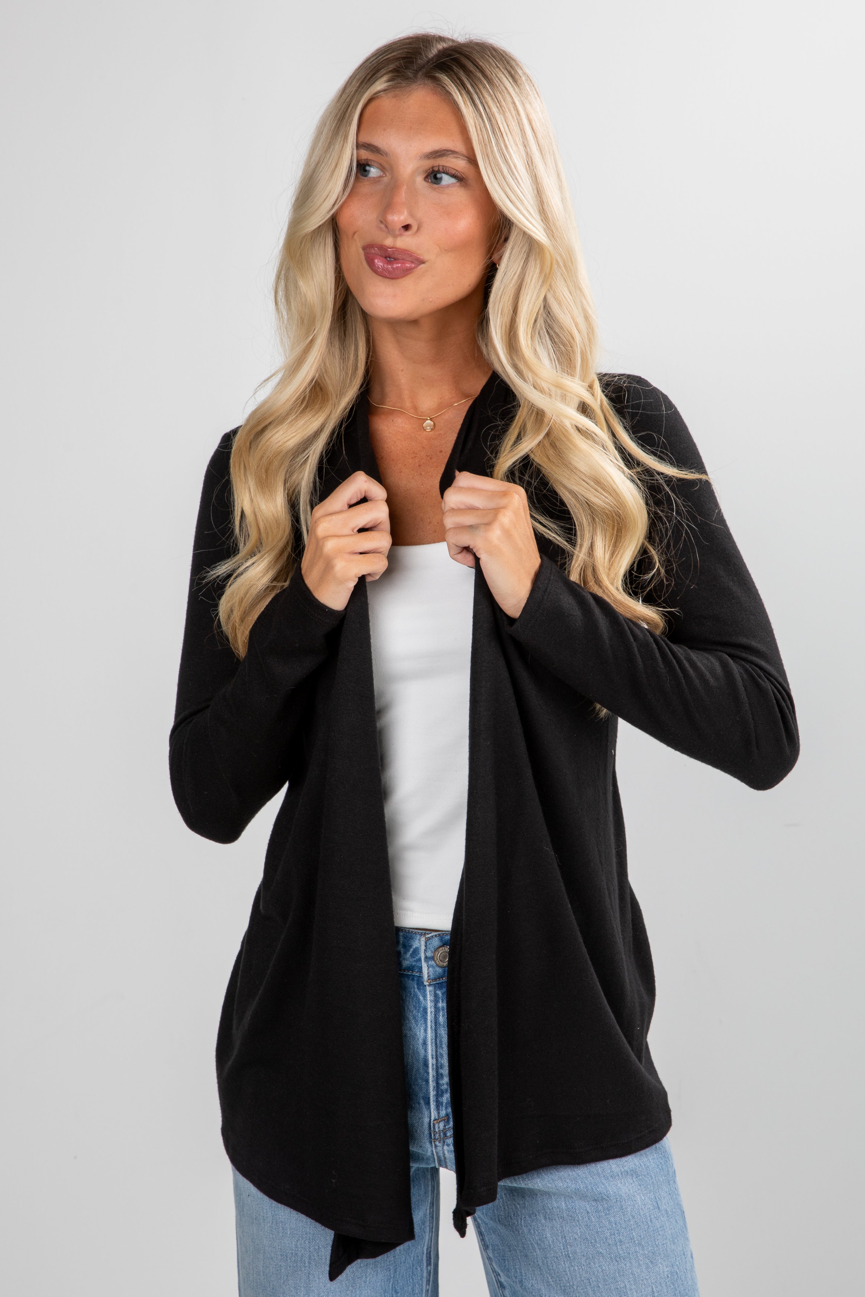 Woman wearing a black cardigan over a white top and blue jeans on a gray background