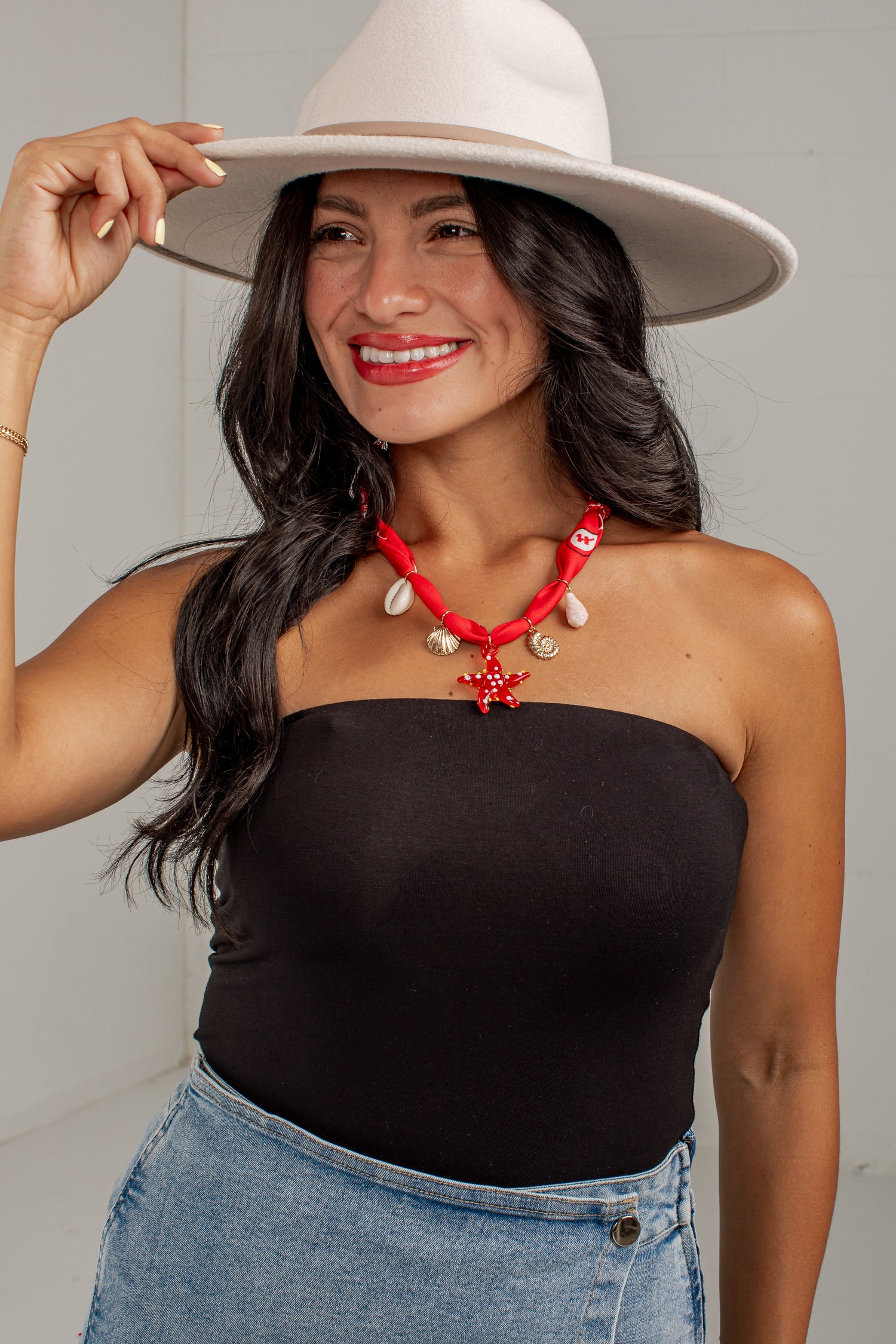 A woman wearing a black strapless tube top, a red and white shell necklace, a white wide-brim hat, and blue denim shorts, posing against a light gray wall.