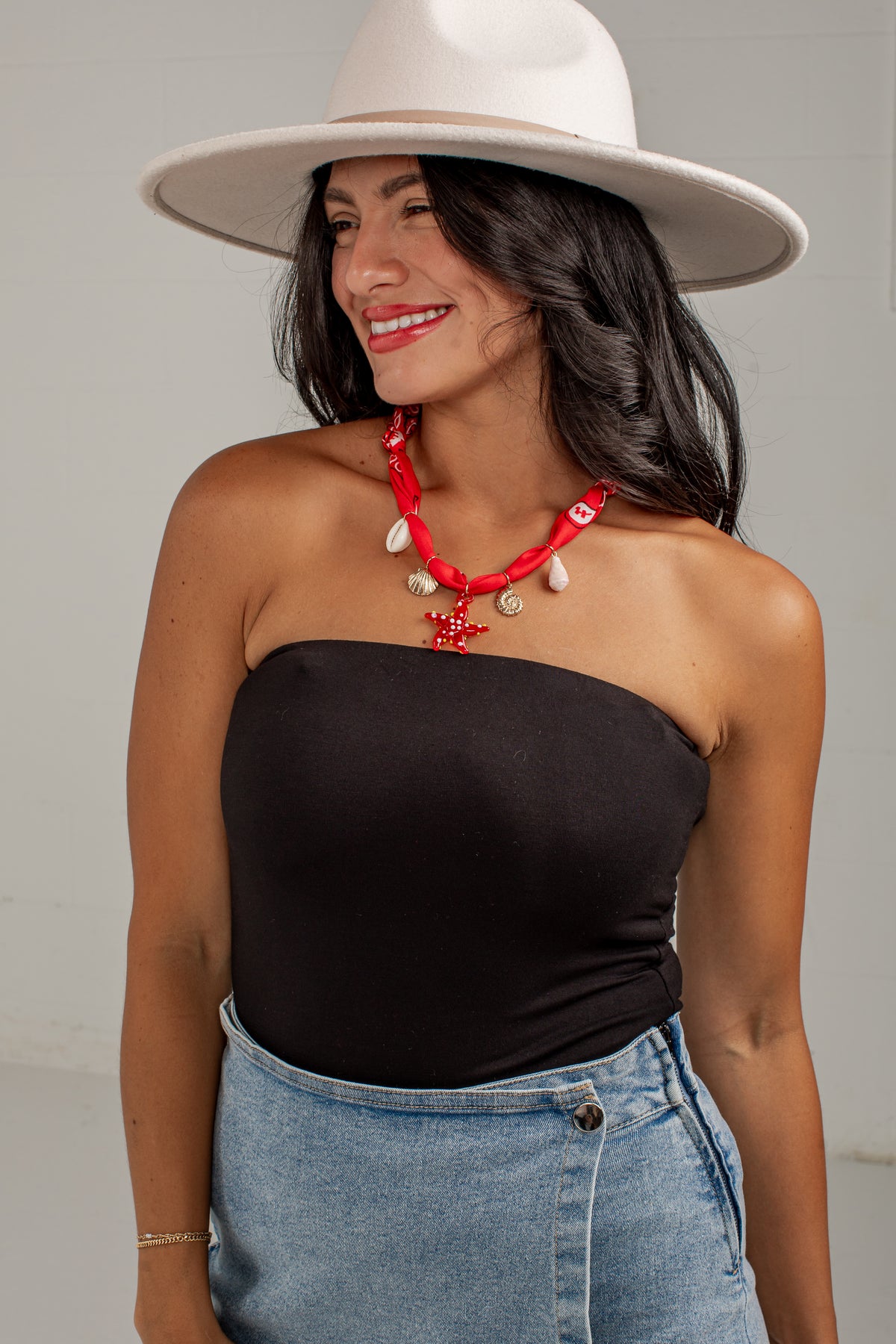 A woman wearing a black strapless bandeau top, light blue denim skirt, white wide-brim hat, and a red beaded necklace with white shell charms, posing against a light gray wall.