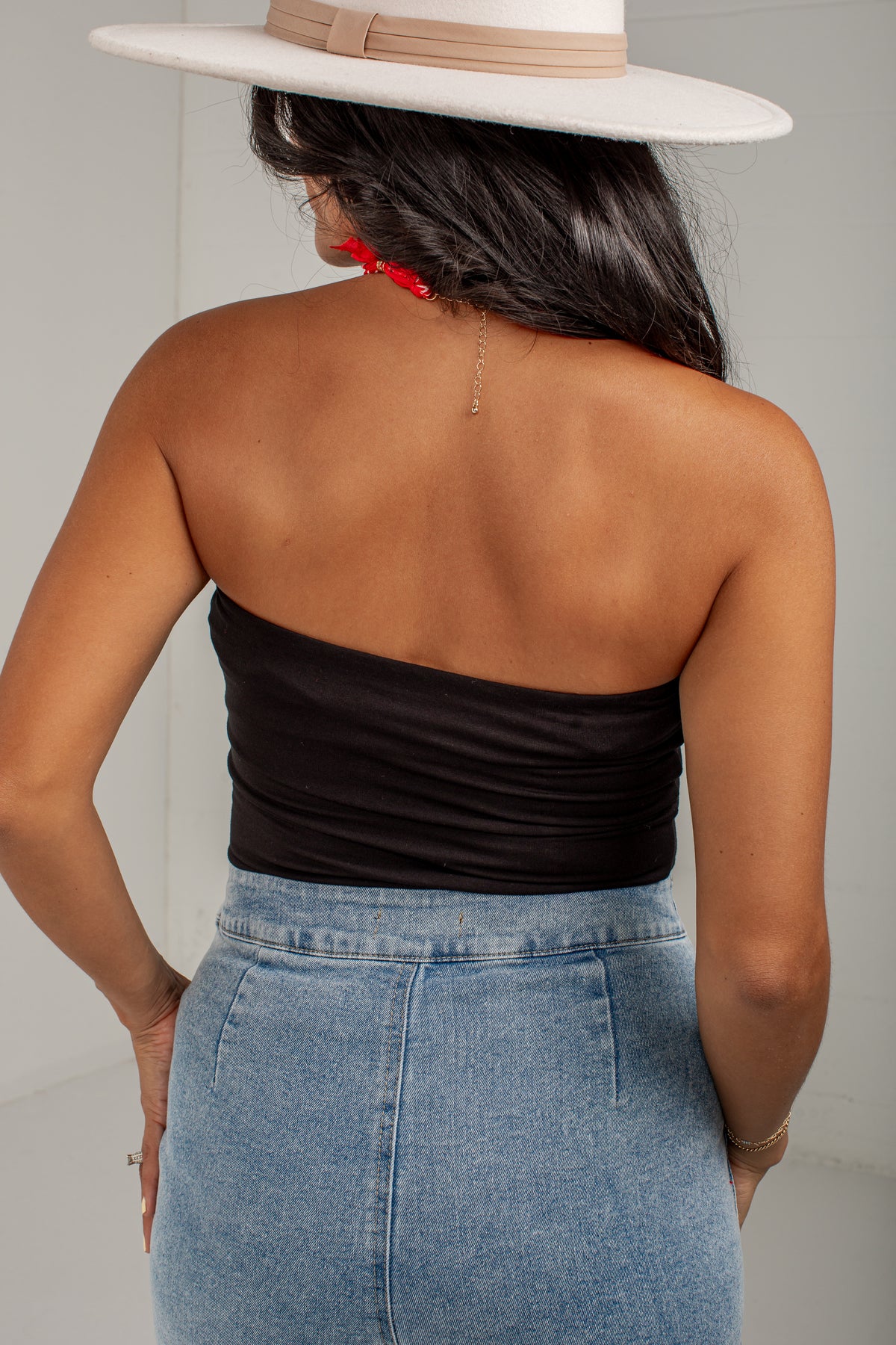 Rear view of a woman wearing a black strapless bandeau top, a light blue denim skirt, and a white wide-brim hat.