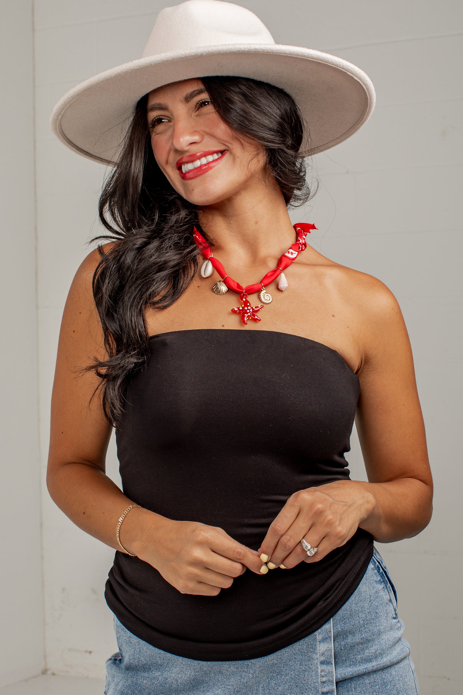 A woman wearing a black strapless tube top, blue jeans, a white wide-brim hat, and a red shell necklace, posing against a light gray wall.