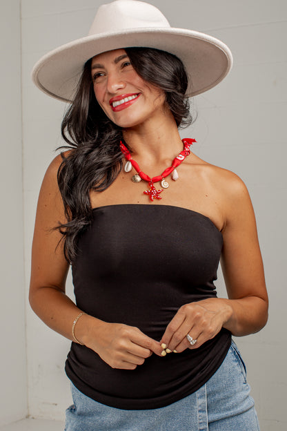 A woman wearing a black strapless tube top, blue jeans, a white wide-brim hat, and a red shell necklace, posing against a light gray wall.