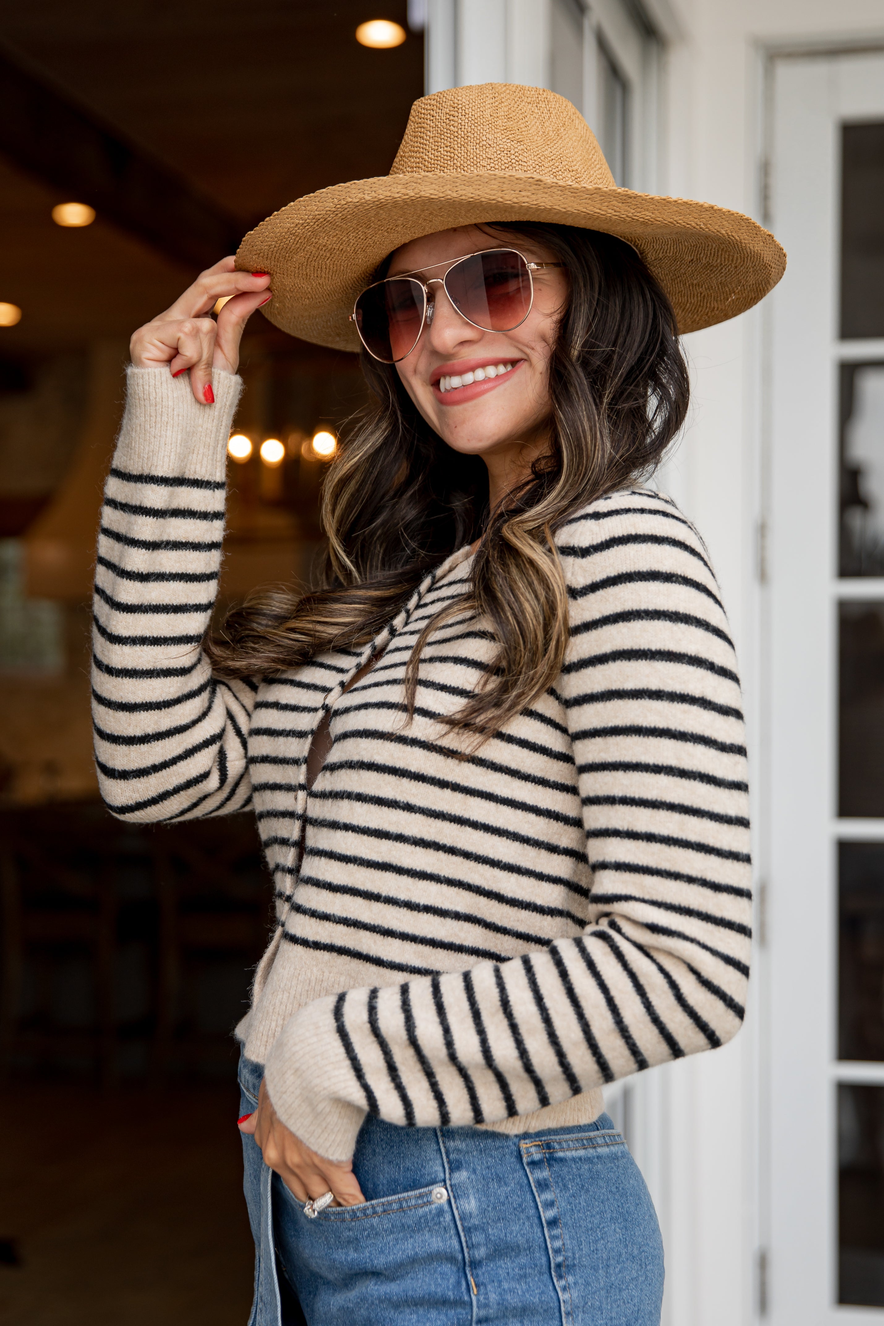Woman wearing a striped sweater, wide-brimmed hat, and sunglasses indoors.