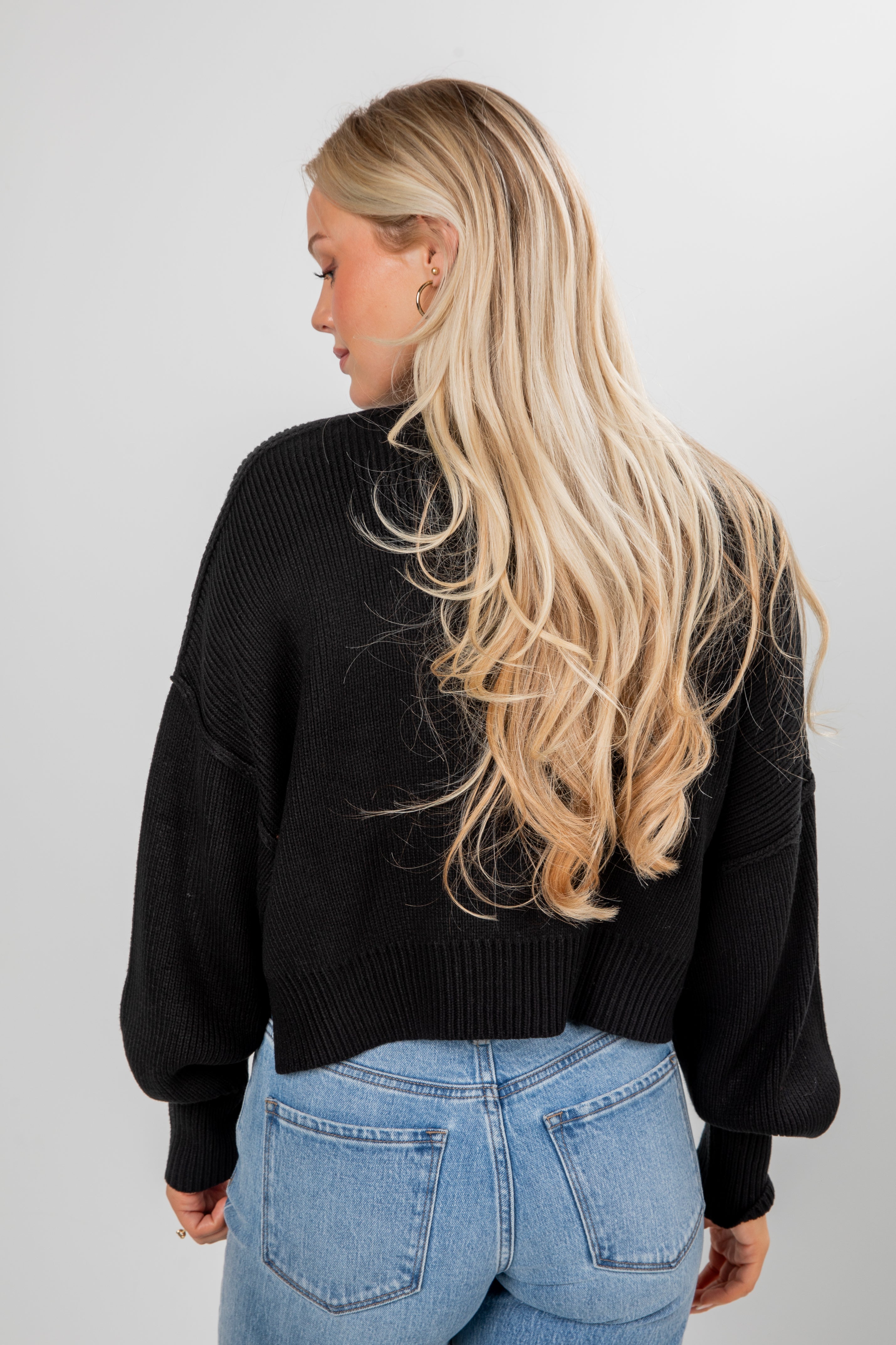Person wearing a black sweater and blue jeans with long blonde hair on a plain background