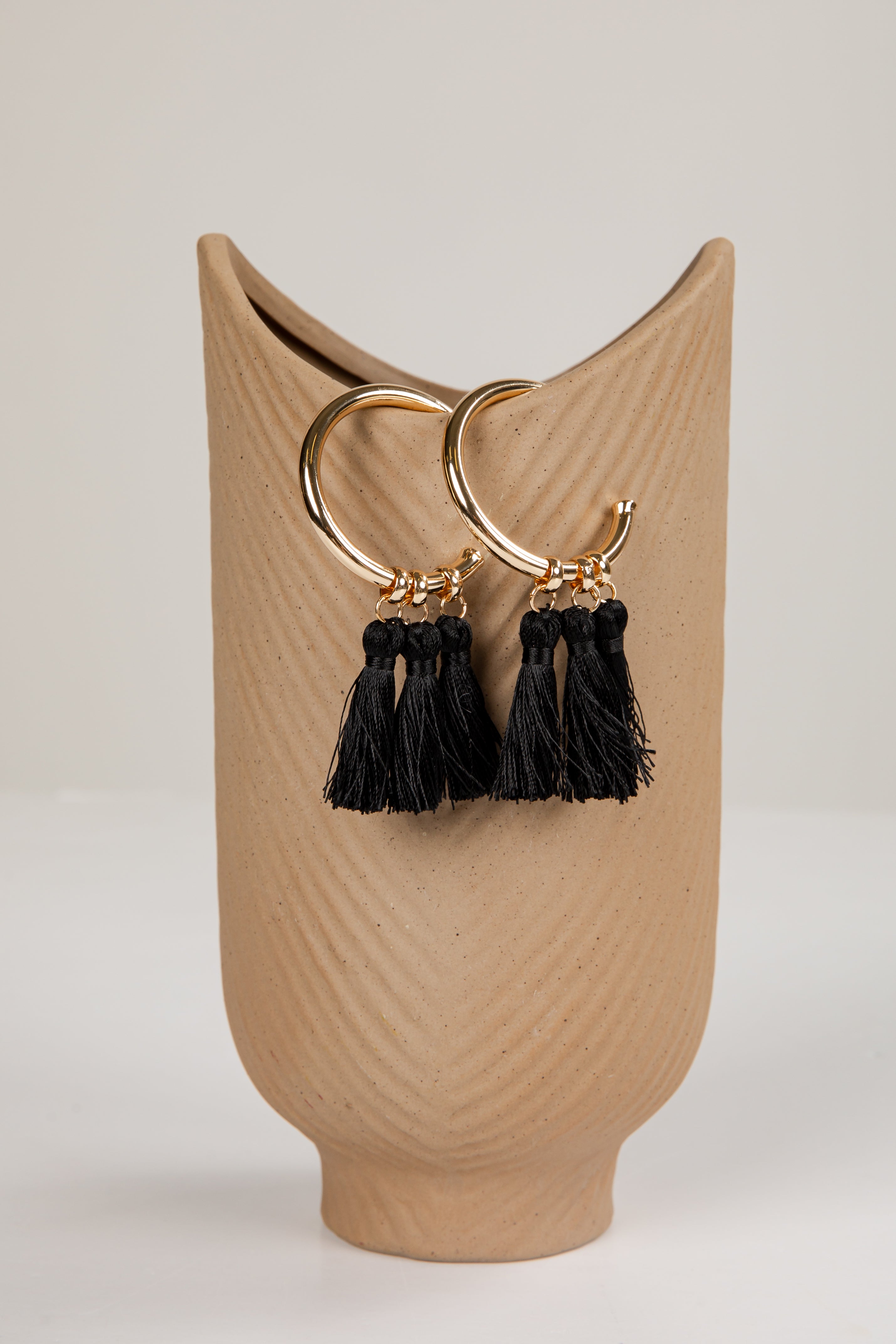 Gold hoop earrings with black tassels on a beige stand against a light background