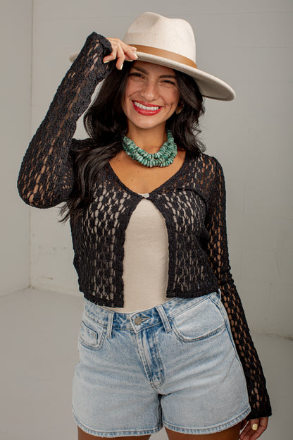 A woman modeling a black long-sleeve lace cardigan over a beige top, paired with light blue denim shorts and a cream fedora hat, against a plain light gray studio background.