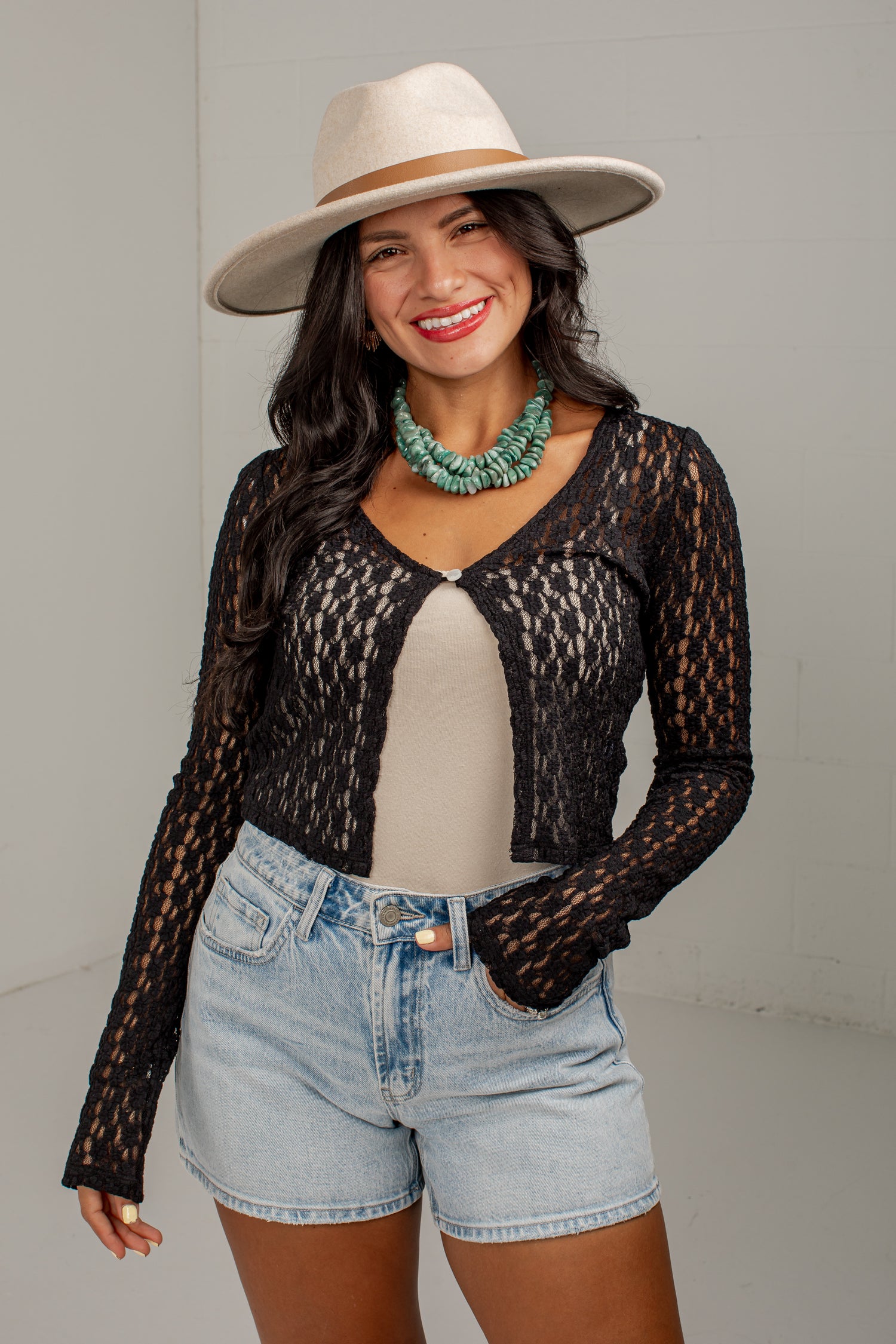 A woman modeling a black long-sleeve lace cardigan over a beige top, paired with light-wash denim shorts and a beige wide-brim hat, against a plain light gray studio background.